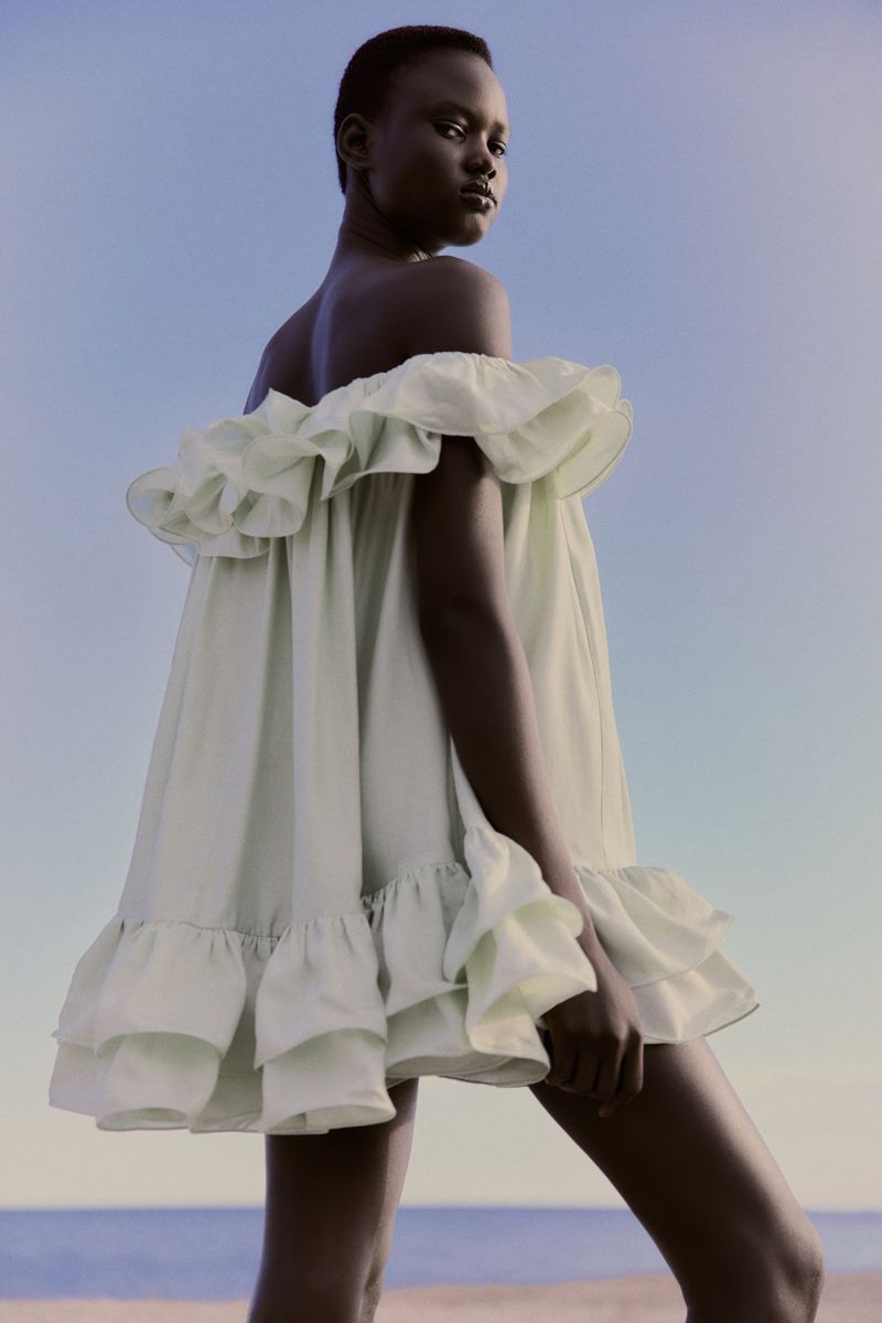 A fashionable Black model poses confidently on a beach in a ruffled white dress against a blue sky, capturing a blend of elegance and serenity.