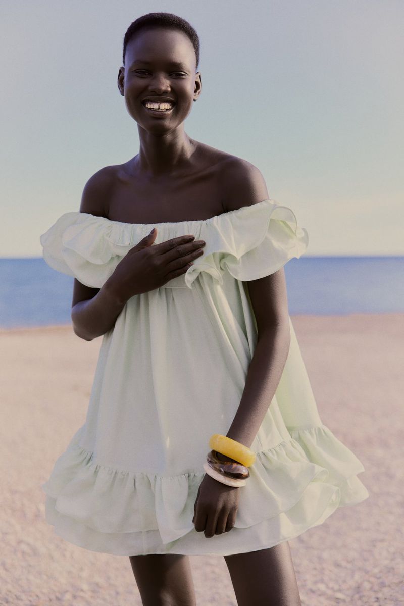 Smiling woman in a light green dress on a beach, accessorized with bracelets, enjoys a sunny day with the ocean in the distance.