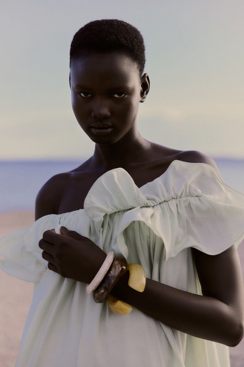 A woman with short dark hair poses in front of a beach in an off-the-shoulder top and bracelets in this portrait shot.