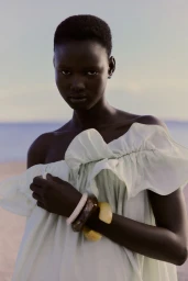 Black woman wearing a light off-shoulder dress and chunky bangles outdoors against a distant ocean horizon.