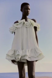 Nyakong Chan wearing a pale green ruffled off-the-shoulder mini dress outdoors against a calm coastal horizon.