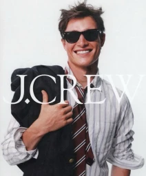 Smiling man wearing sunglasses, striped shirt, and striped tie while holding a dark blazer over his shoulder for J.Crew.