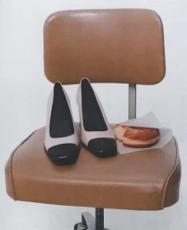 Two-tone pumps and a frosted pastry on a paper wrapper resting on a brown office chair against a white background.