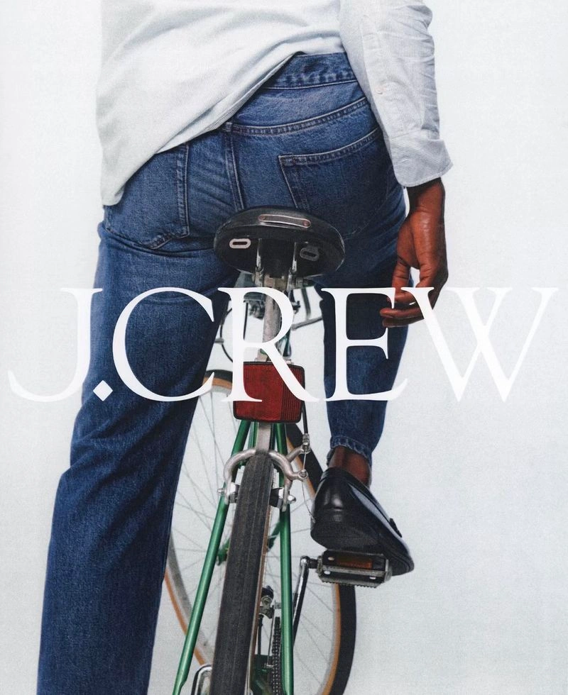 Person straddling a green bicycle wearing blue jeans, a white long-sleeve shirt, and black loafers against a white backdrop.