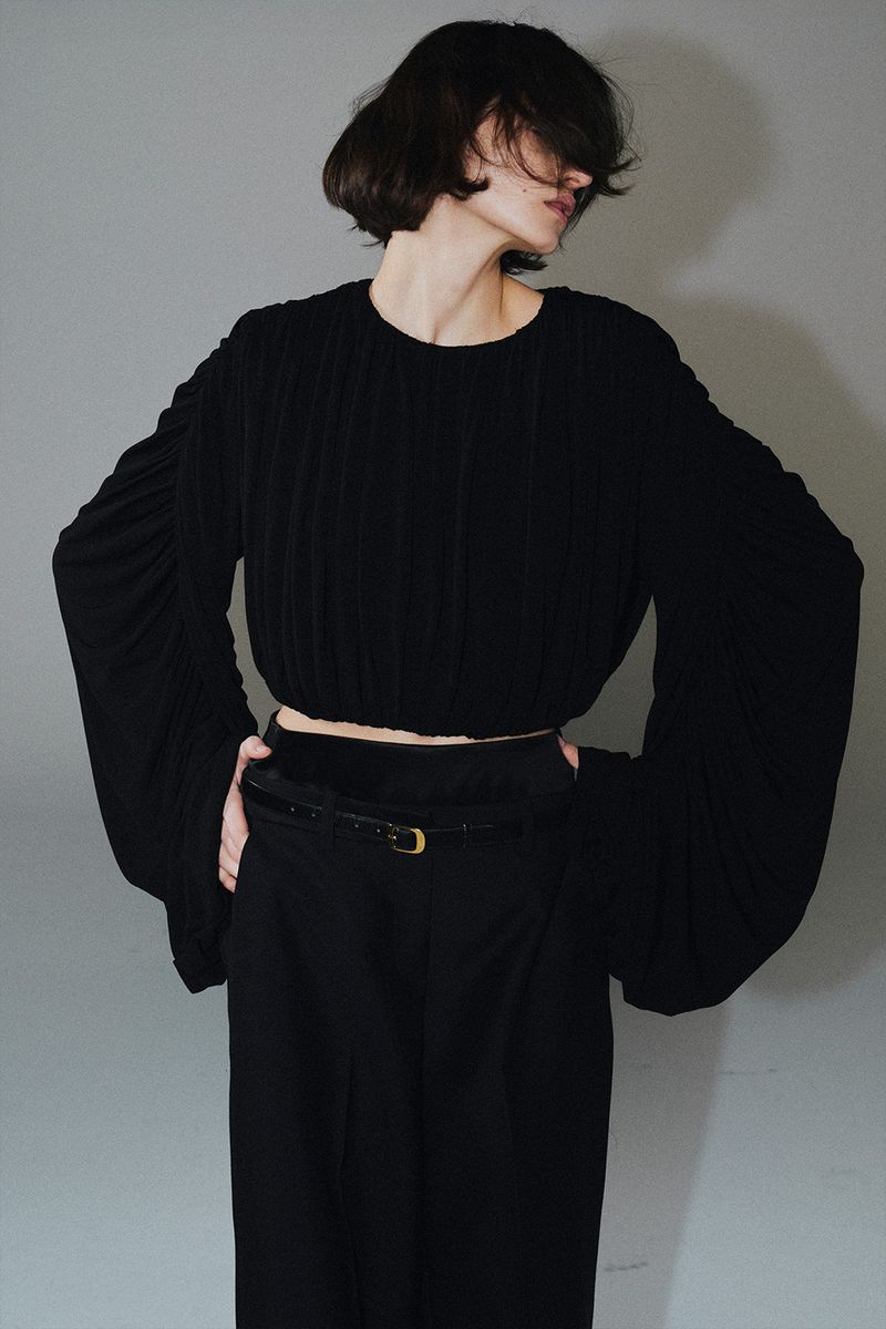 Model with dark hair wearing a black long-sleeve top and trousers against a grey seamless background