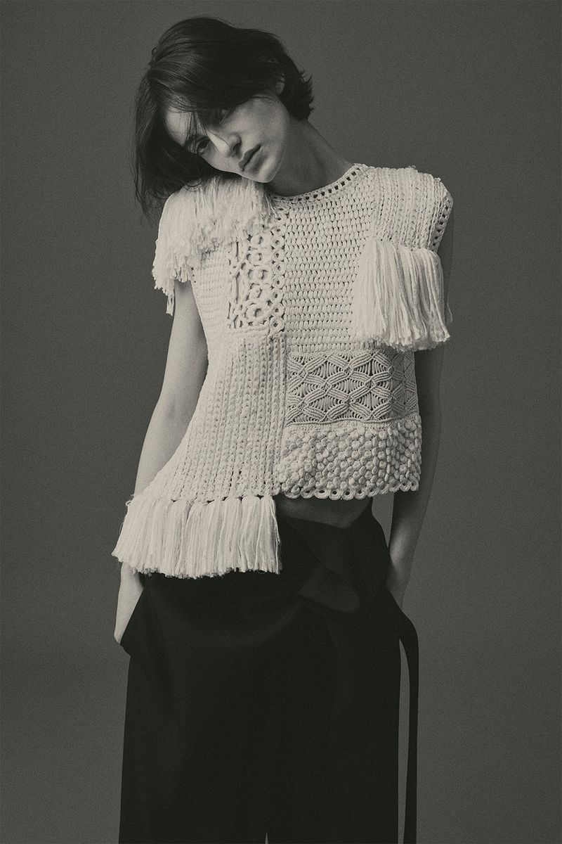 Model with dark hair wearing a white crochet top and dark trousers against a grey backdrop