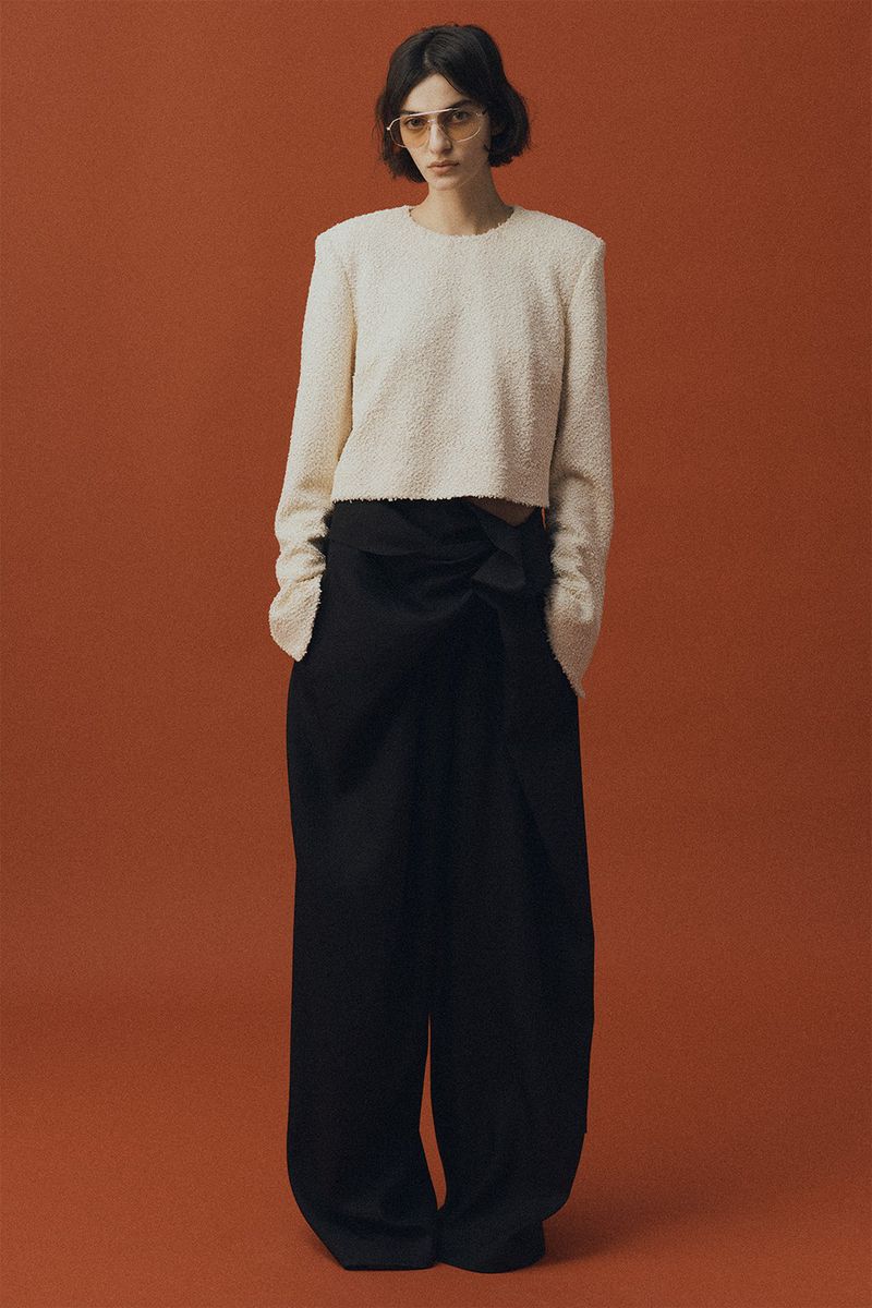 Model wearing a textured cream top and wide black trousers against a warm terracotta background