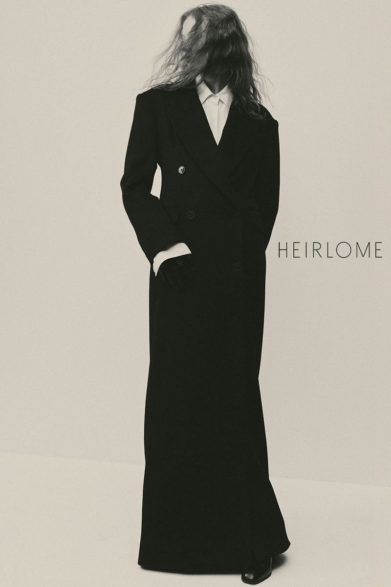 Model with long tousled hair wearing a long black coat over a white collared shirt against a neutral background