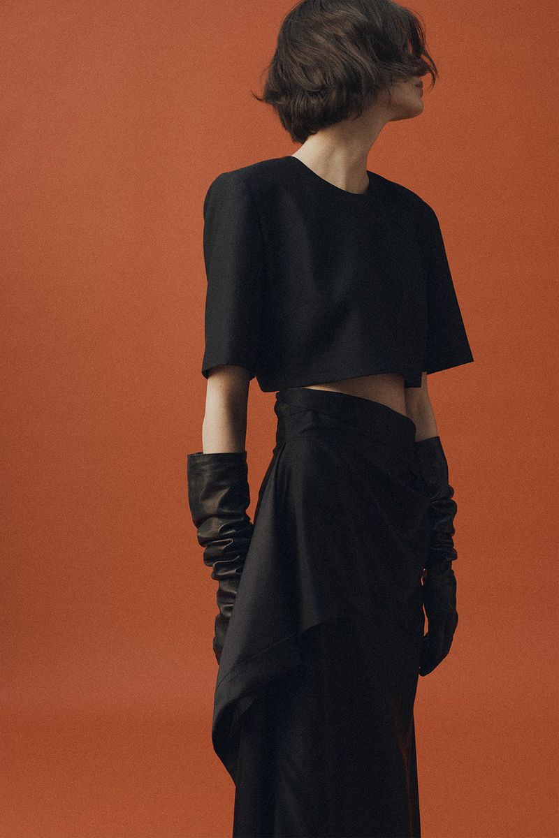 Model in black short-sleeve top and skirt wearing long leather gloves against a rust orange background.