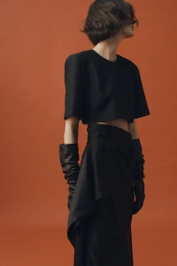 Model in black short-sleeve top and skirt wearing long leather gloves against a rust orange background.
