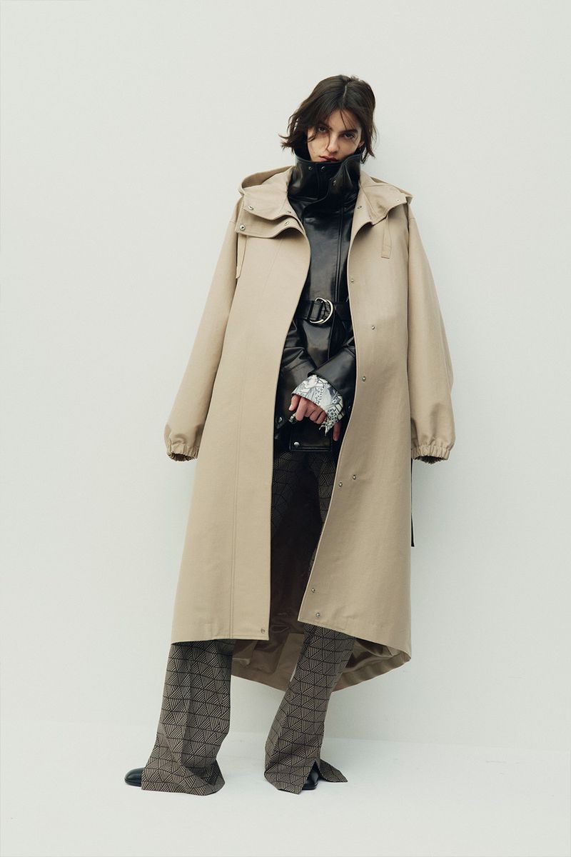 Model wearing a long beige hooded coat over a black jacket and patterned trousers against a white background