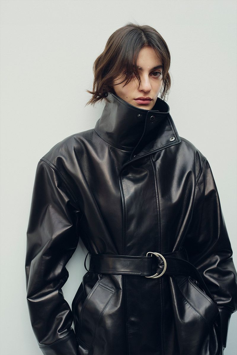 Model in oversized black leather jacket with high collar and belted waist against white background
