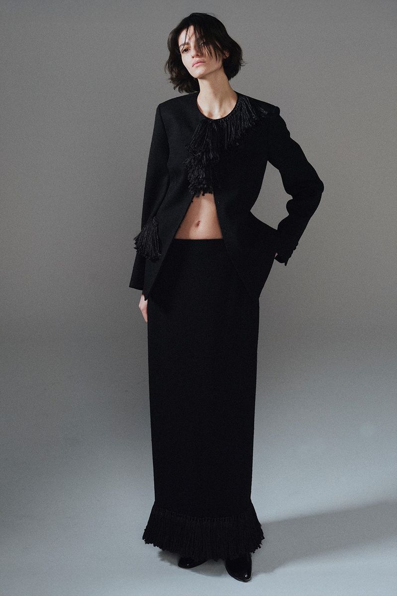Model wearing a black jacket with fringed detailing and a matching long skirt against a plain grey seamless background.