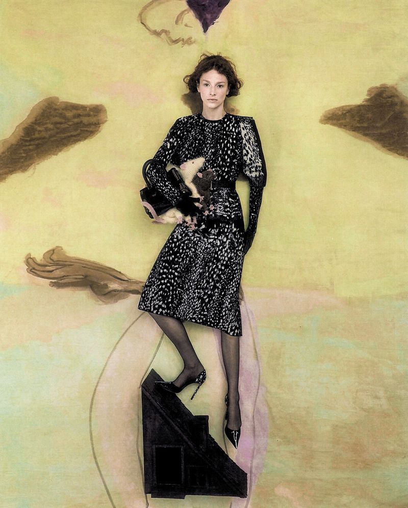 Hunter Schafer wearing a black patterned dress standing on a small dark triangular structure against a painted textured background