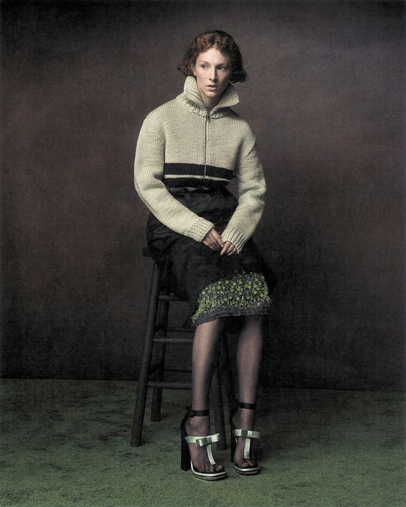 Hunter Schafer seated on a wooden stool wearing a cream zip-up knit sweater and black skirt in a studio portrait.