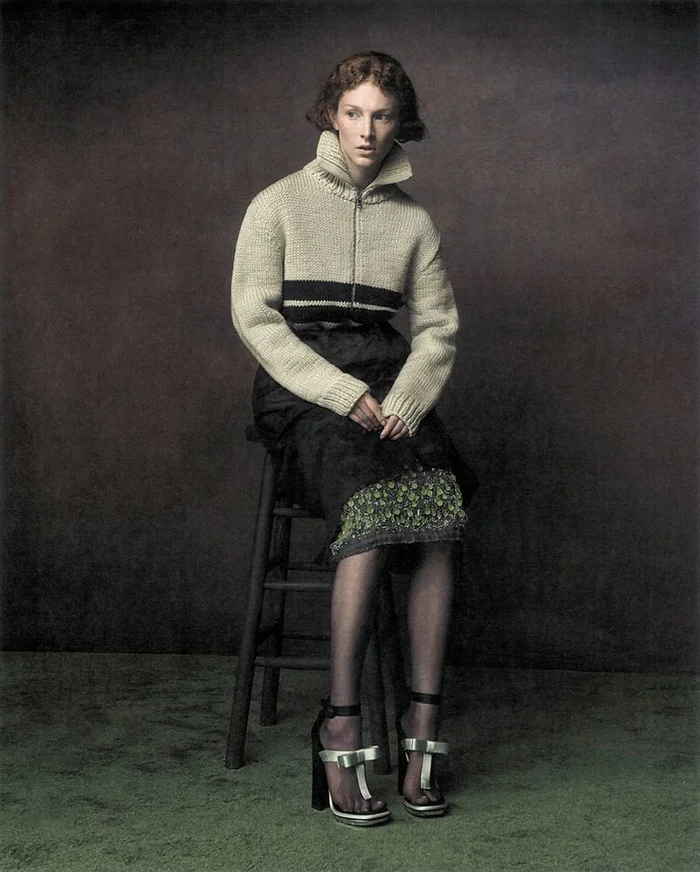 Hunter Schafer seated on a wooden stool wearing a cream zip-up knit sweater and black skirt in a studio portrait.