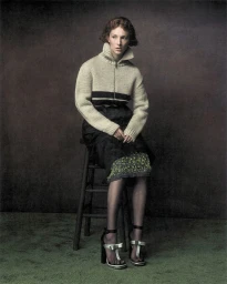 Hunter Schafer seated on a wooden stool wearing a cream zip-up knit sweater and black skirt in a studio portrait.