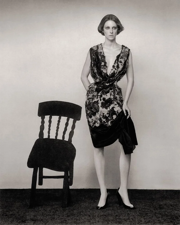Hunter Schafer wearing a patterned dress standing next to a dark wooden chair against a plain studio background.