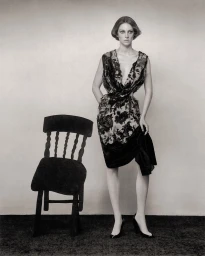 Hunter Schafer wearing a patterned dress standing next to a dark wooden chair against a plain studio background.