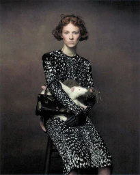 Hunter Schafer seated wearing a patterned black and white dress holding a handbag and stuffed animals