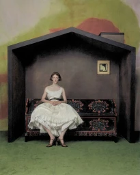 Hunter Schafer seated inside a dark house-shaped pavilion wearing a white dress on a painted bench