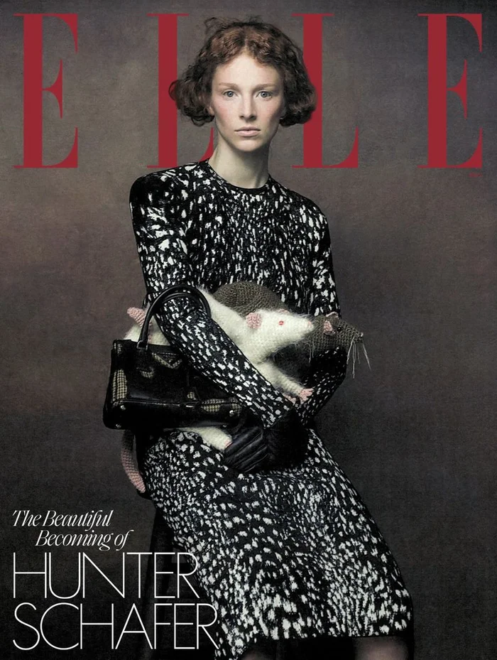 Hunter Schafer seated wearing a black and white patterned dress holding a toy rat and a black handbag
