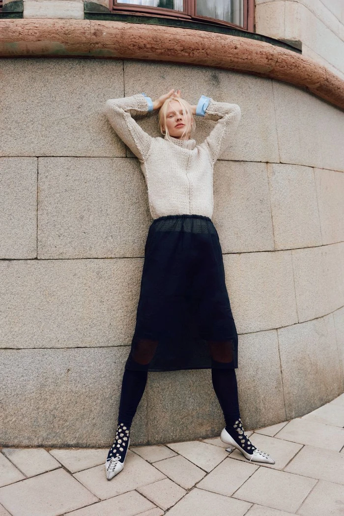 Model Vilma Sjöberg leaning against a curved stone wall in a cream textured sweater and dark midi skirt for Elle Sweden.