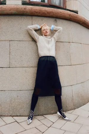 Model Vilma Sjöberg leaning against a curved stone wall in a cream textured sweater and dark midi skirt for Elle Sweden.
