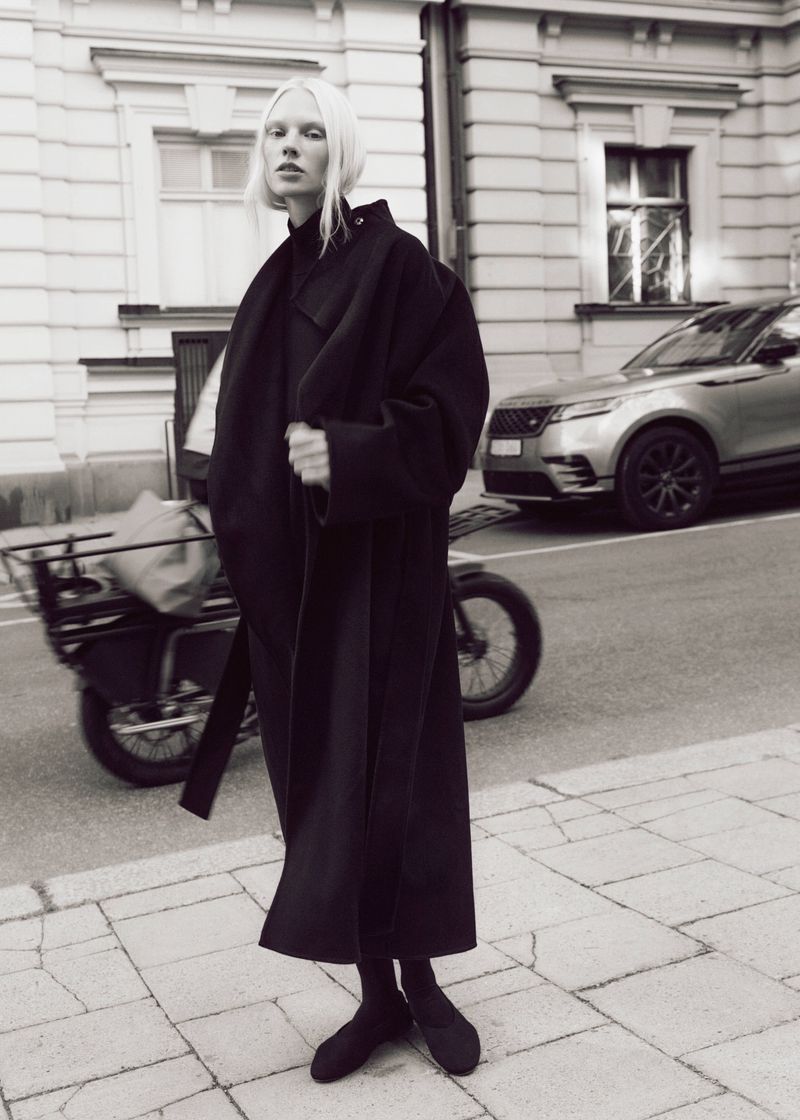 Model with blonde hair wearing a long black coat standing on a city street with a parked car and bicycle in the background