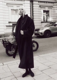 Model with blonde hair wearing a long black coat standing on a city street with a parked car and bicycle in the background