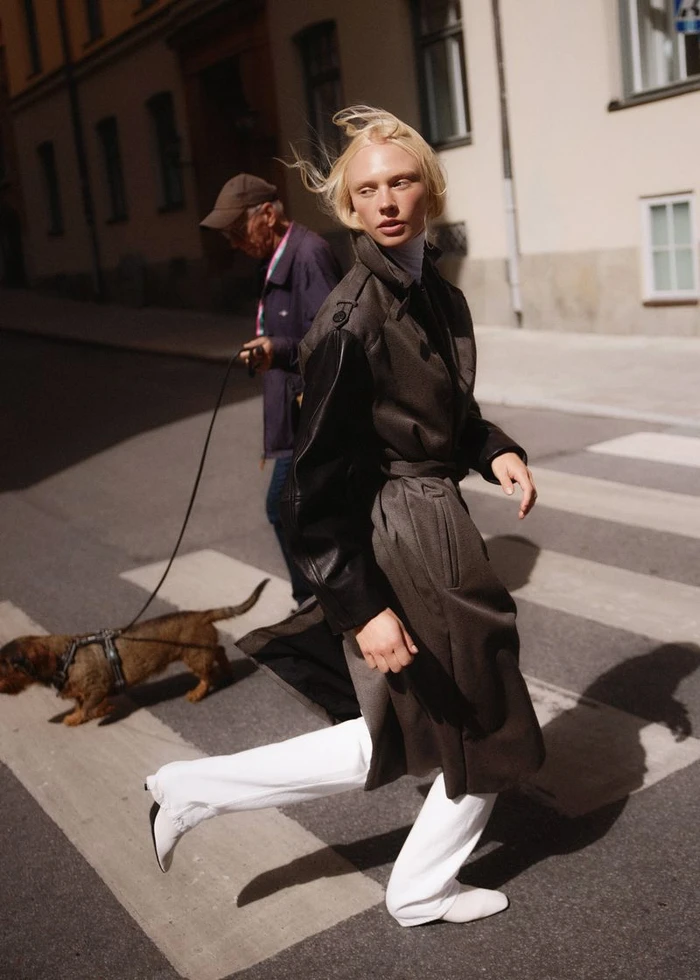 Blonde model in a dark coat and white trousers walking across a city street with a small dog and handler