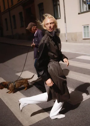 Blonde model in a dark coat and white trousers walking across a city street with a small dog and handler