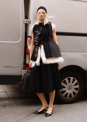 Model Vilma Sjoberg wearing a textured black vest and black skirt standing beside a silver van for Elle Sweden.