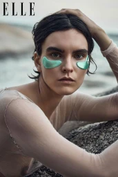 Model wearing green under-eye patches and sheer long-sleeve top outdoors near rocky coast for Elle Bulgaria