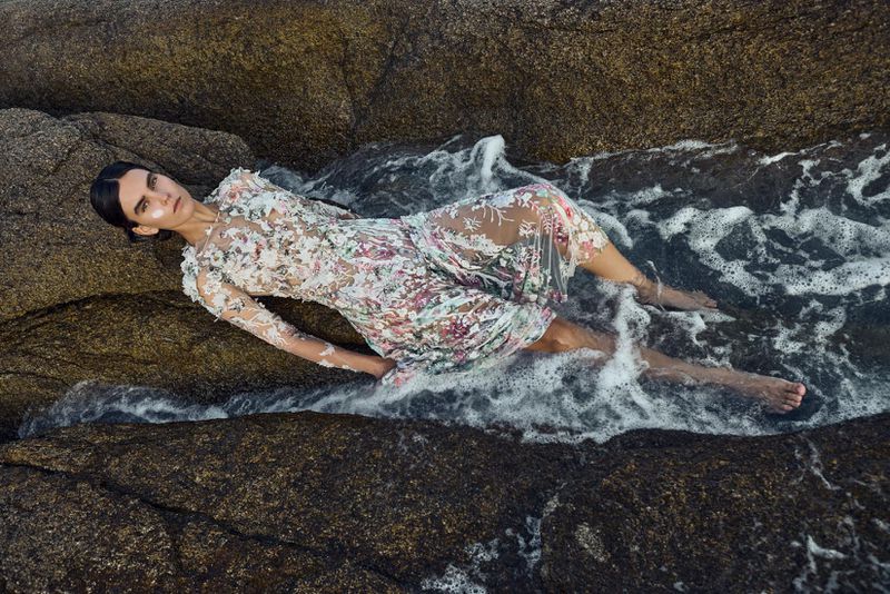 Model lying on wet rocky shore in a sheer floral embroidered dress surrounded by splashing ocean water
