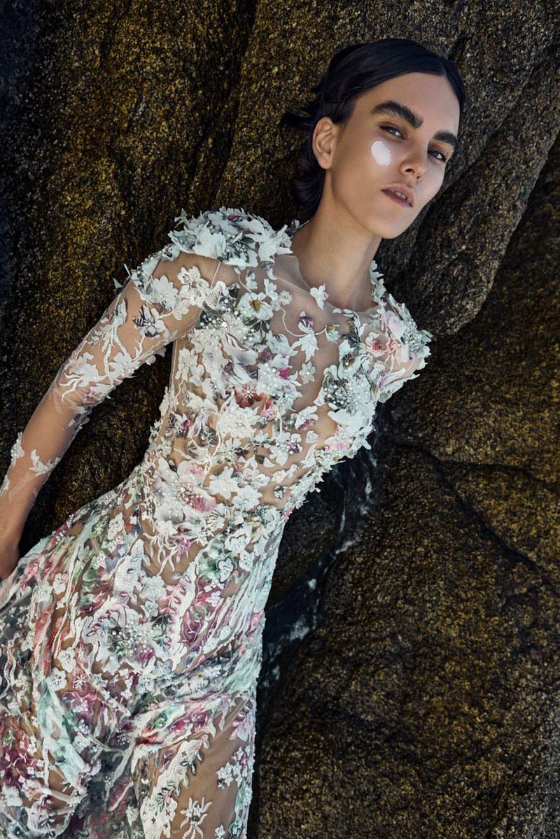 Model in intricate floral embroidered sheer gown reclining against dark rock surface