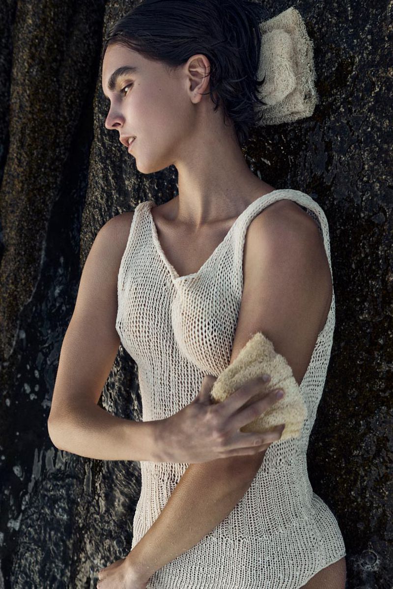 Model in a cream knitted tank top exfoliating her arm with a loofah against a dark rocky coastal background