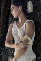Model in a cream knitted tank top exfoliating her arm with a loofah against a dark rocky coastal background