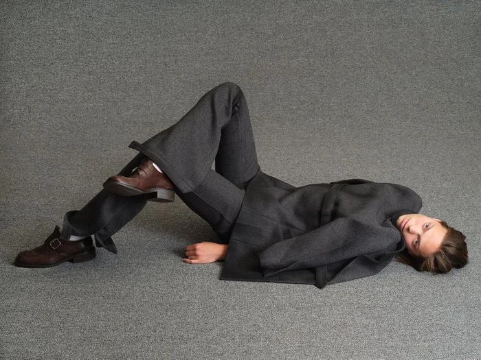 Model lying on a textured grey carpet wearing a dark grey suit and brown leather shoes for Family Style.
