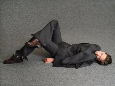 Model lying on a textured grey carpet wearing a dark grey suit and brown leather shoes for Family Style.
