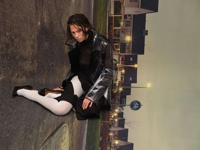 Model wearing a translucent vinyl coat, black turtleneck, white tights, and black heels sitting on an asphalt street