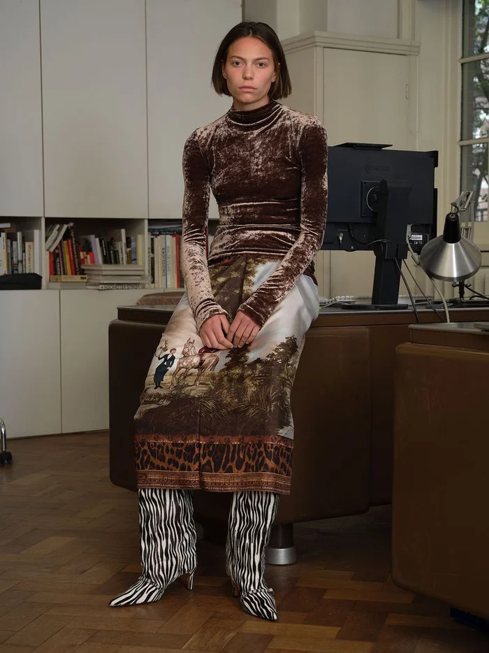 Model seated on a desk in an office wearing a brown velvet top, a printed patterned skirt, and zebra print boots.