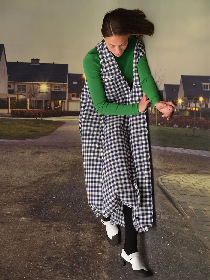 Model in green top and blue and white checkered garment posing against a backdrop of a suburban street.