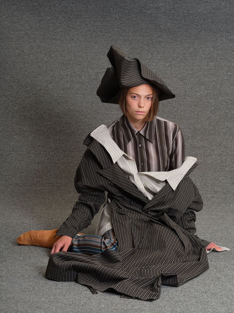 Model seated on grey textured floor wearing layered striped garments and an asymmetrical sculptural hat.
