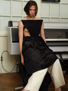 Model with dark hair wearing a black textured dress and wide cream trousers seated in an interior room