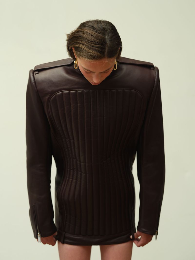 Model wearing a structured dark brown leather jacket with prominent shoulder pads against a seamless backdrop