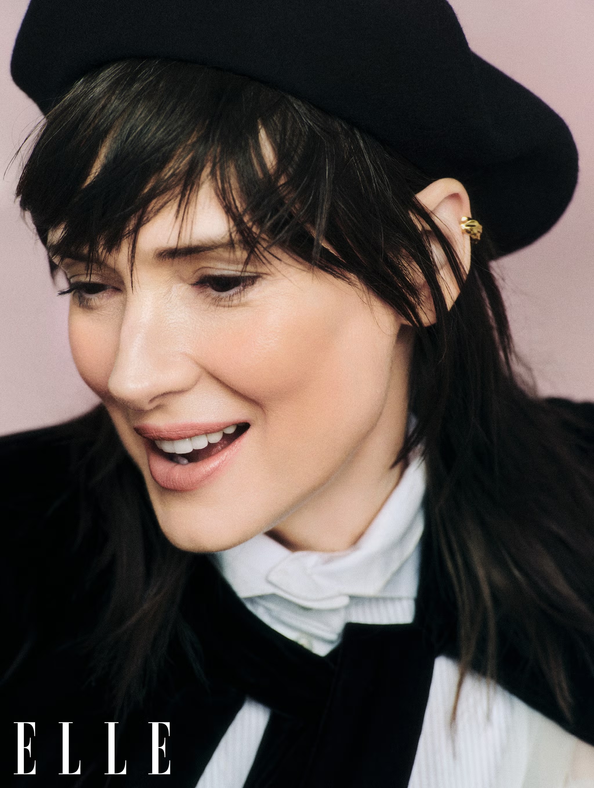 Portrait of a woman with dark hair wearing a black hat and velvet jacket. Smiling with an ear cuff, shot in a pink-toned studio.
