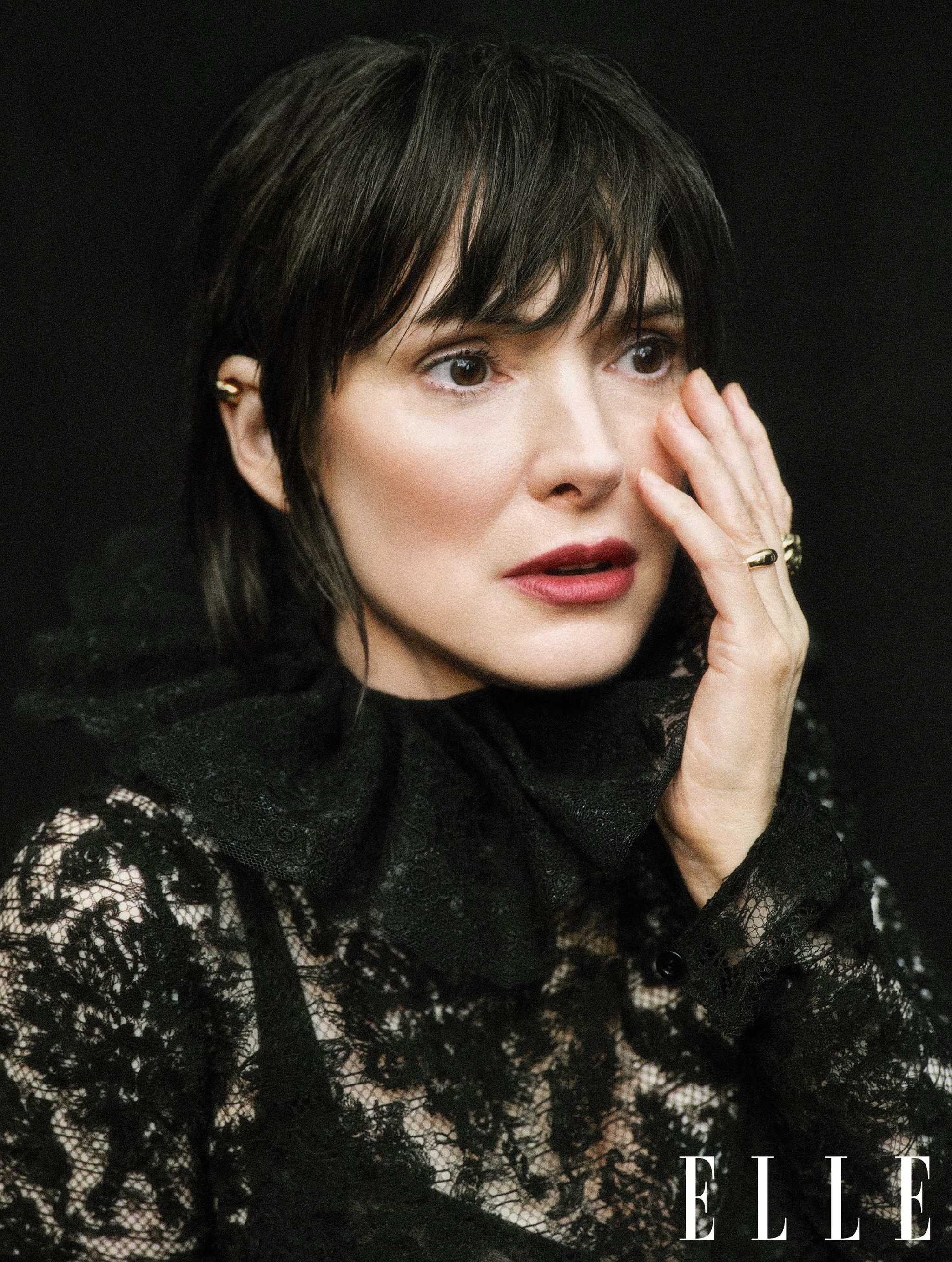 Close-up portrait of a woman with dark hair and fair skin wearing a black lace top and gold earrings in a dark photo studio.