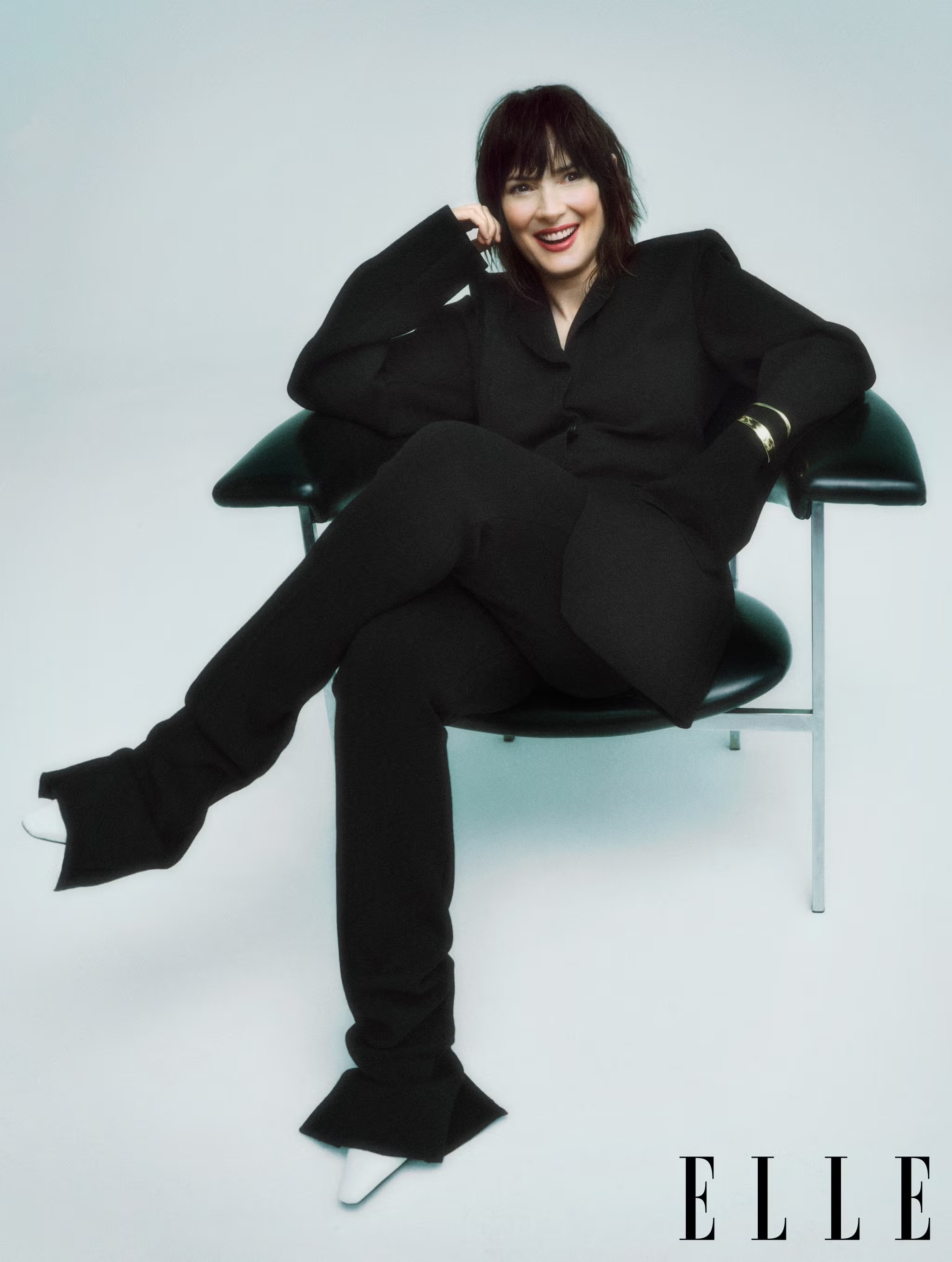 Elegant Winona Ryder seated in a black suit and white shoes against a bright background in a studio portrait for ELLE magazine.