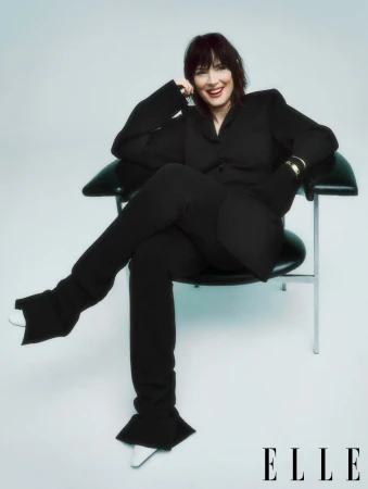 Elegant Winona Ryder seated in a black suit and white shoes against a bright background in a studio portrait for ELLE magazine.